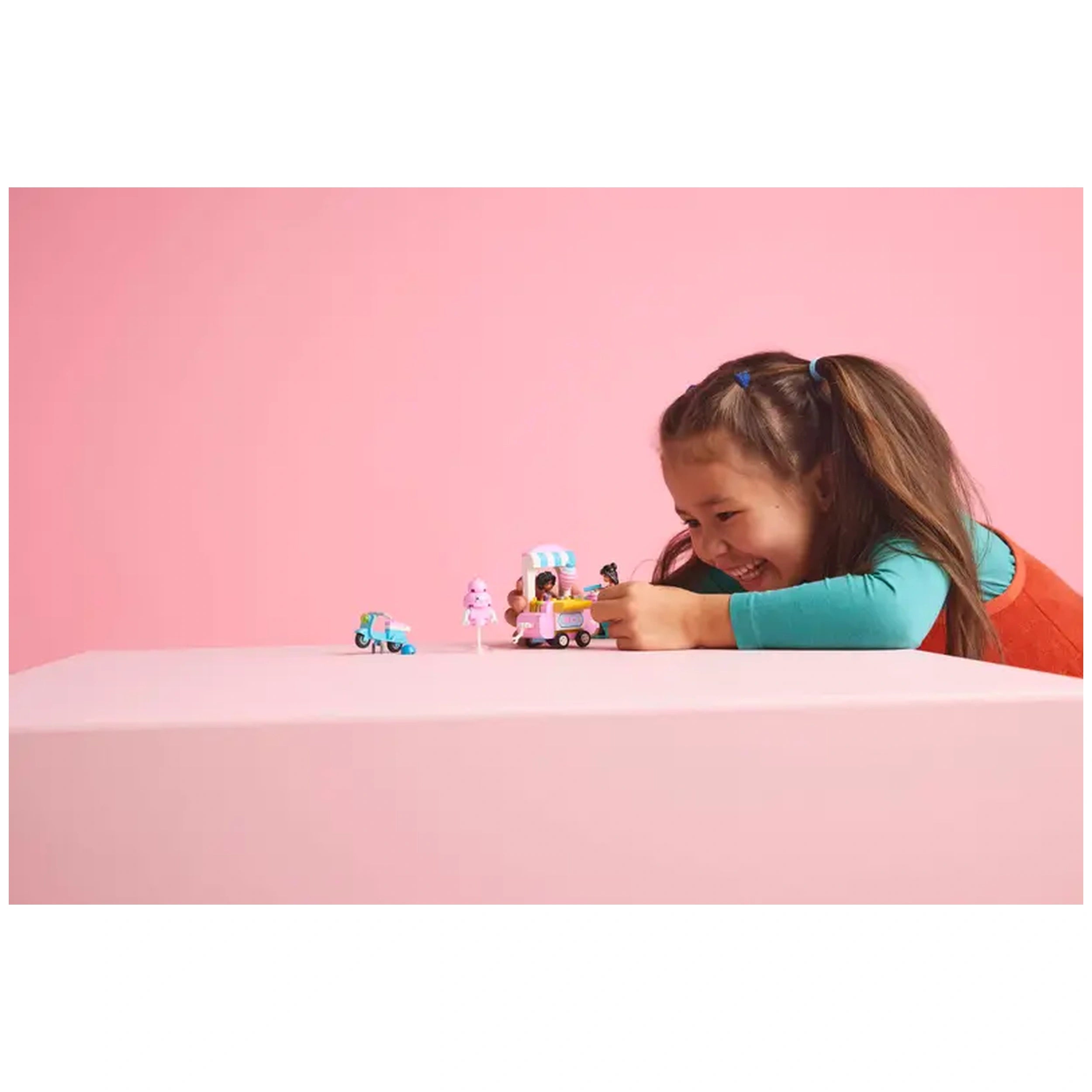 A joyful child interacts with the LEGO Friends Cotton Candy Stand and Scooter set, featuring vibrant toys and two minidolls, on a pastel pink background.
