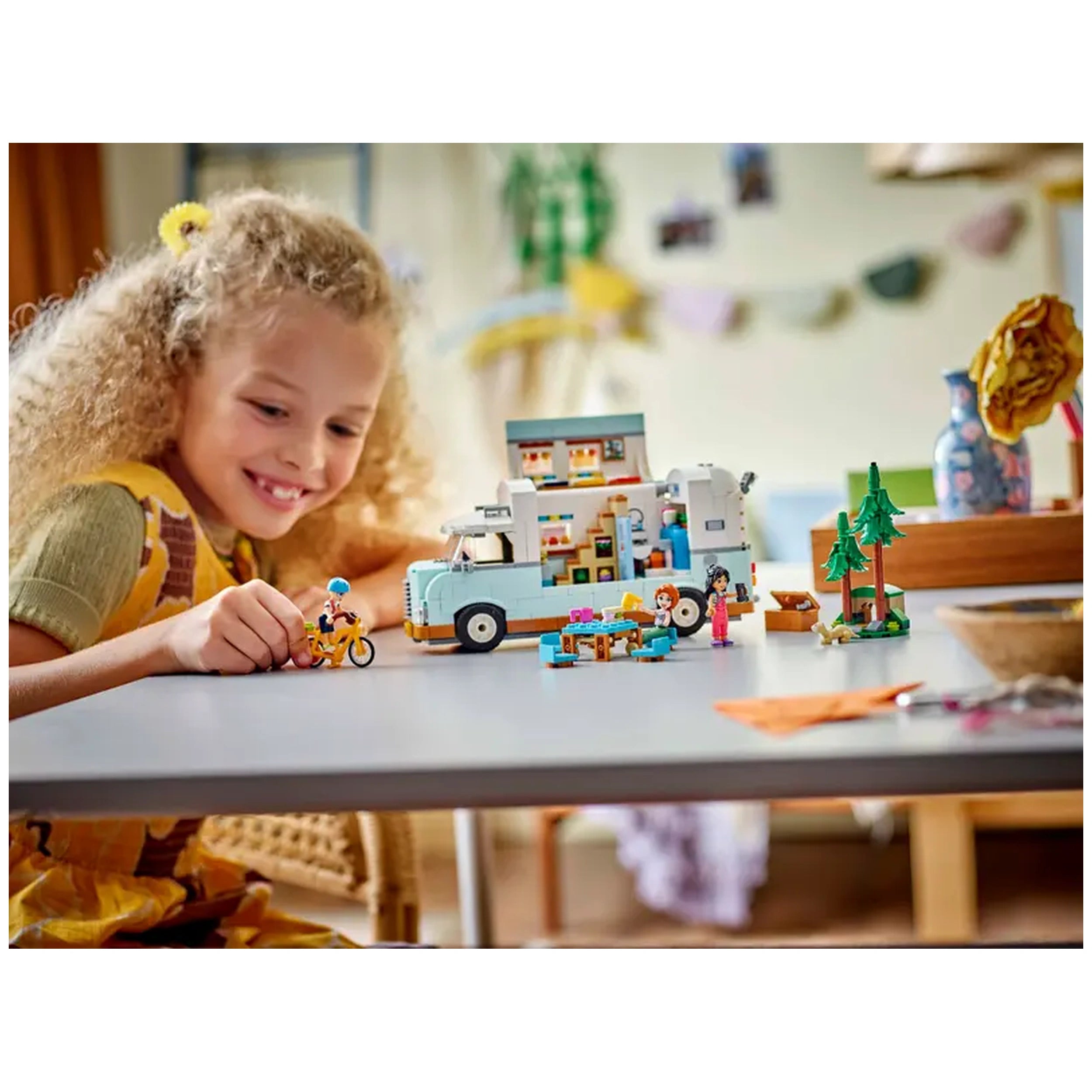 Young girl with curly hair smiling while playing with LEGO® Friends Friendship Camper Van Adventure set, featuring a camper van, mini dolls, and accessories on a table.