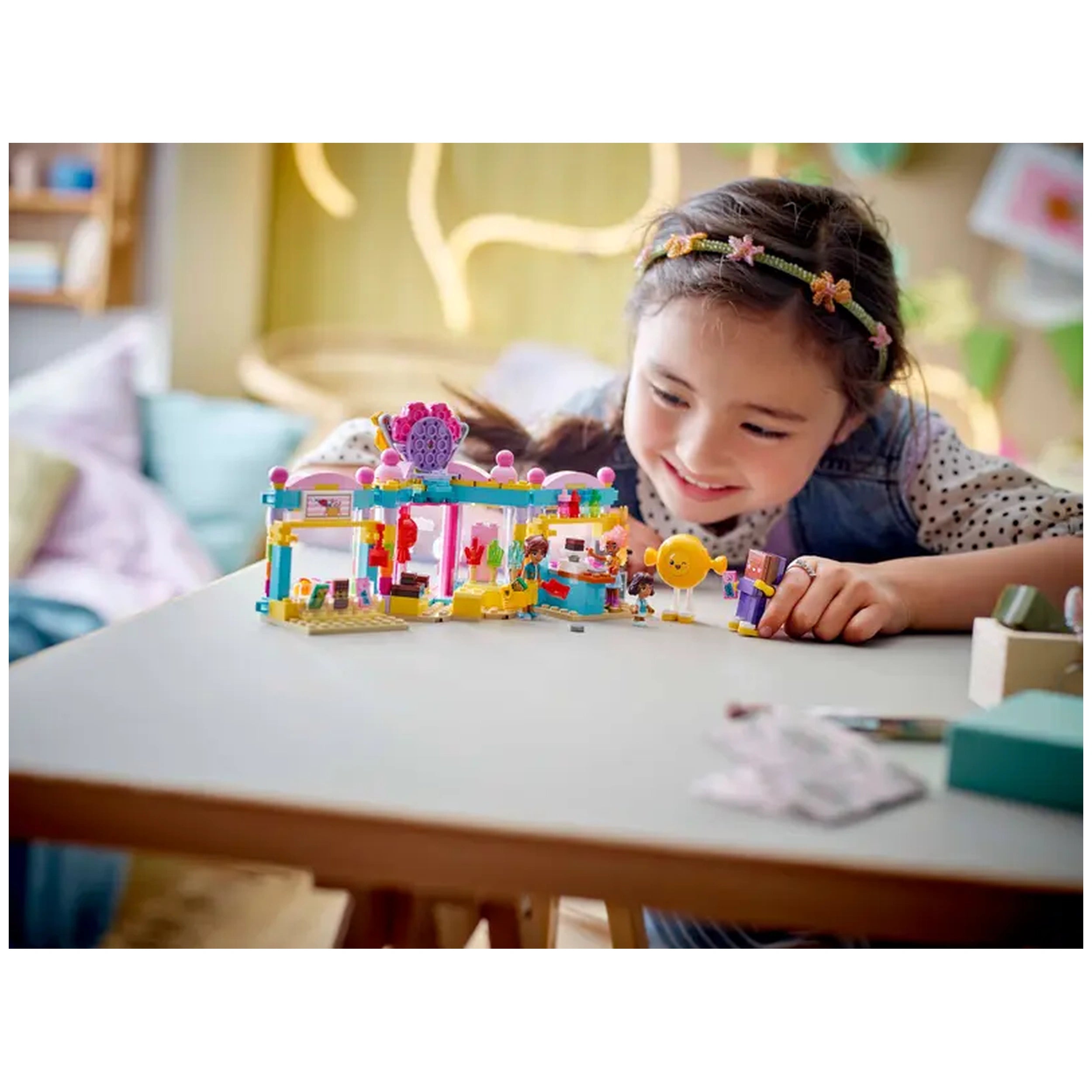 A smiling girl plays with the Lego Friends Heartlake City Candy Store set, featuring colorful storefront and mini-dolls on a light table.