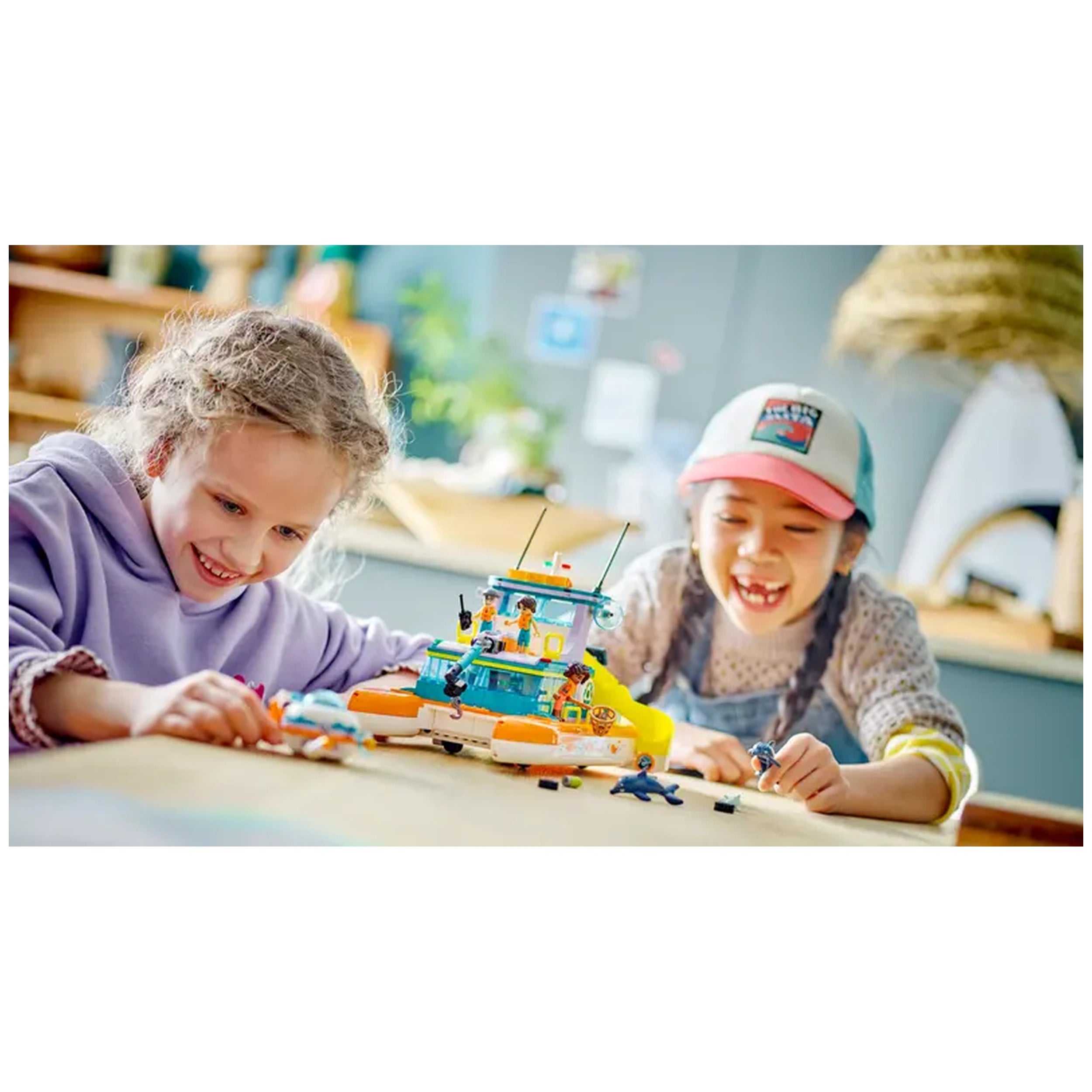 Two smiling girls play with the LEGO® Friends Sea Rescue Boat set on a wooden table, exploring colorful pieces for imaginative ocean adventures.