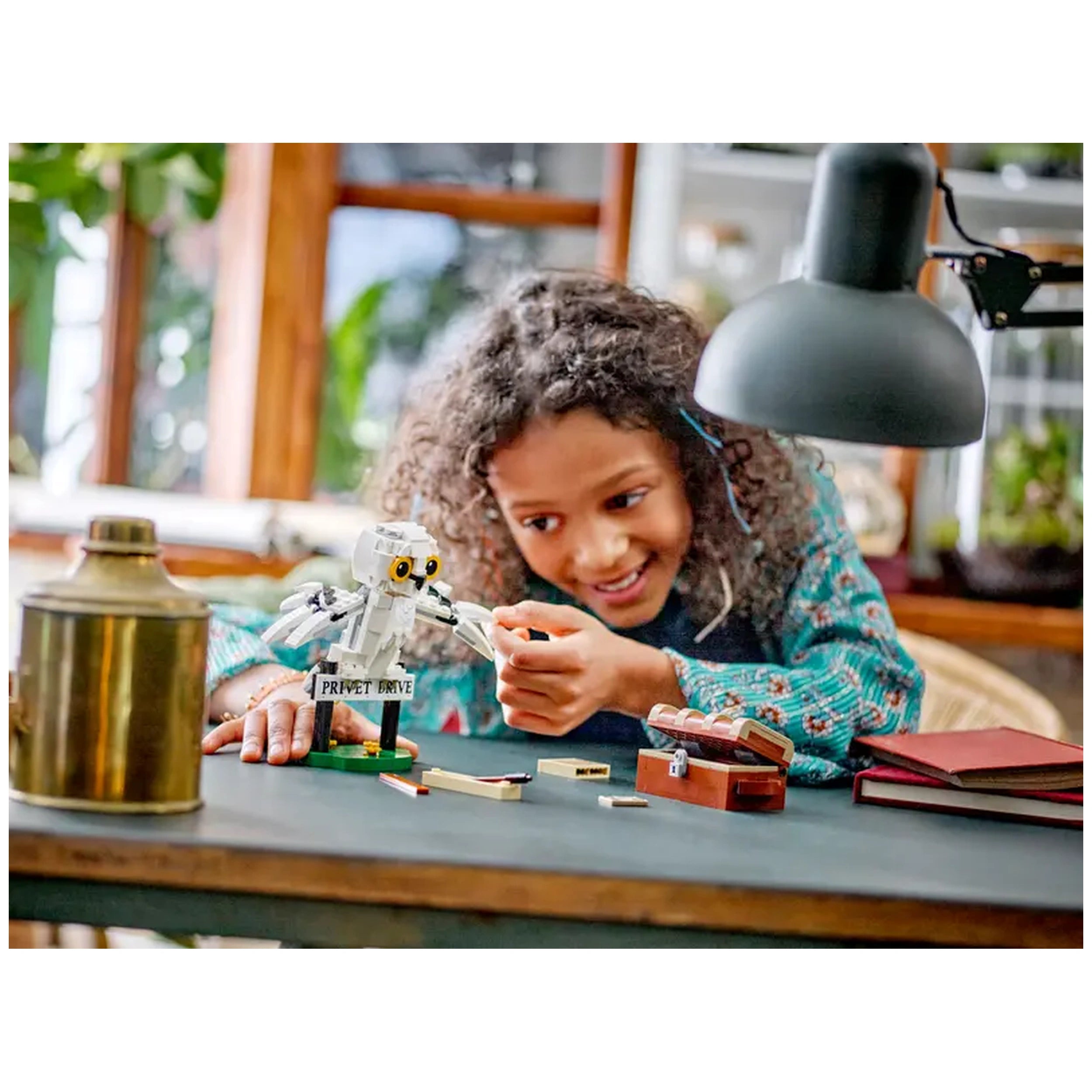 A child with curly hair joyfully assembles the LEGO Harry Potter Hedwig model at a table, featuring a 'Privet Drive' sign, under a lamp.