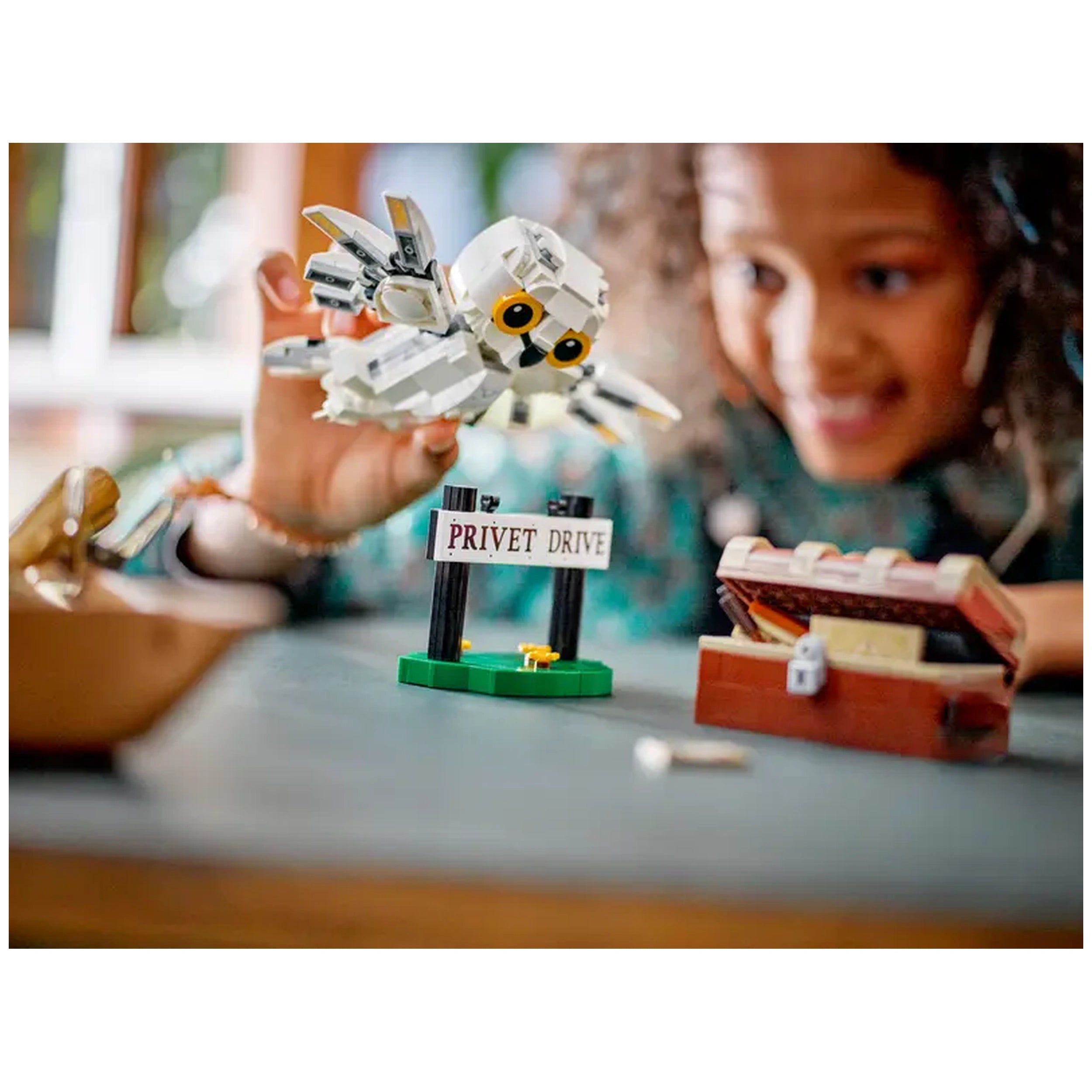 A young child holds a posable Hedwig owl above a green LEGO 'Privet Drive' sign, with a treasure chest on the table.