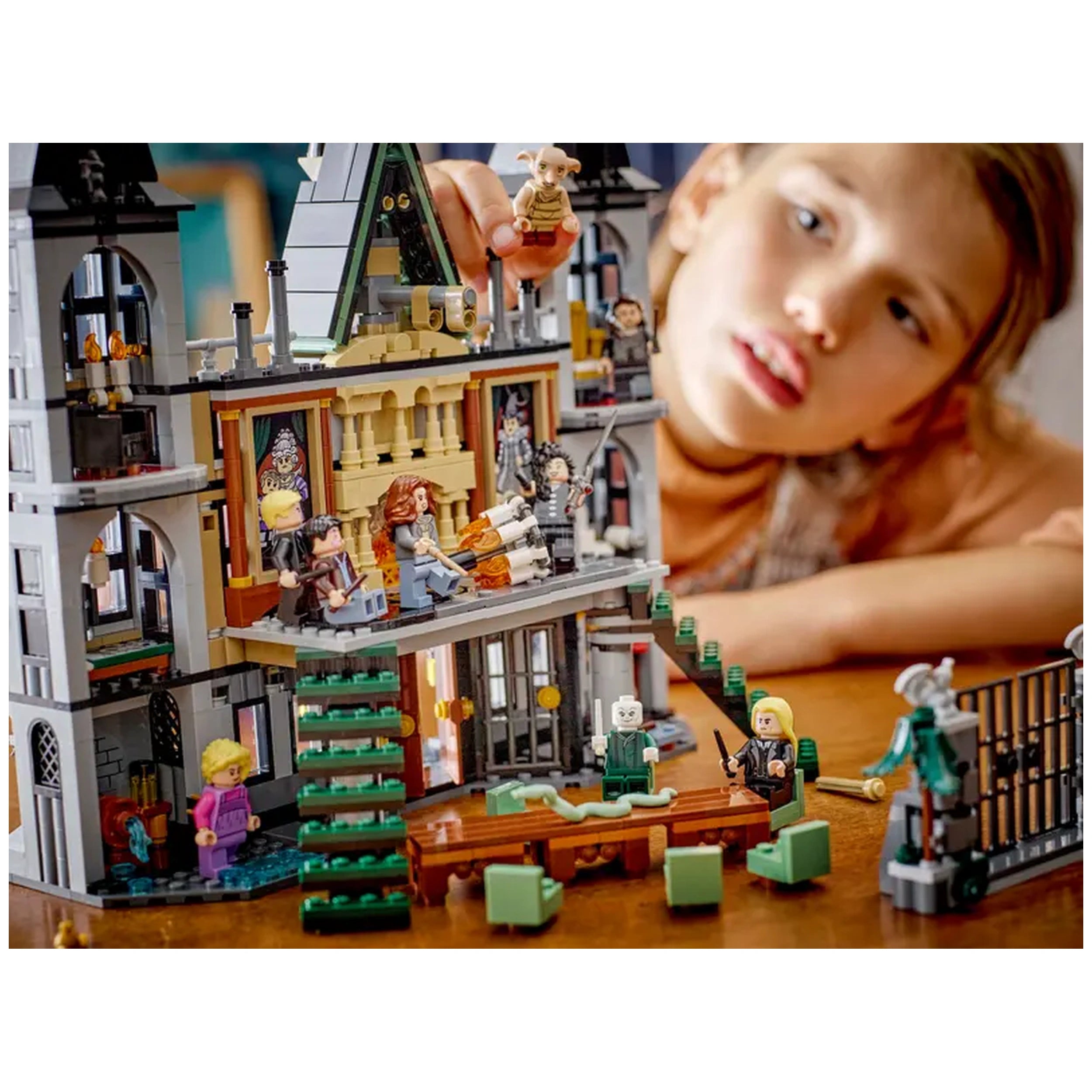 A young child focuses intently on the LEGO Harry Potter Malfoy Manor set, featuring intricate details like figures of Draco Malfoy and Dobby, a green staircase, and scenes from the Wizarding World. The model showcases the manor’s architectural features, including windows and a table, all on a wooden surface.