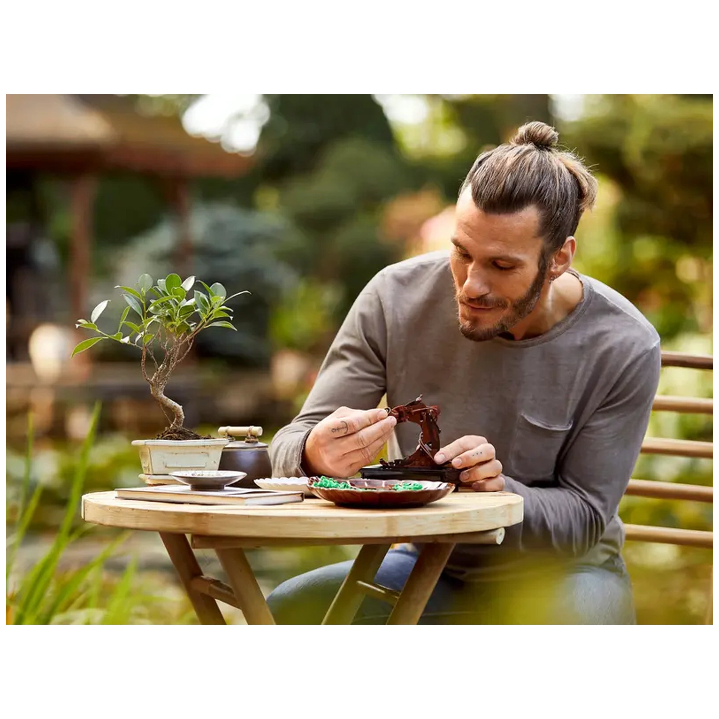 A man with long hair in a bun sits at a wooden table, carefully assembling a LEGO® Bonsai Tree. The small bonsai plant and construction pieces are in front of him, surrounded by greenery. The setting is serene, evoking a tranquil atmosphere ideal for creative expression.
