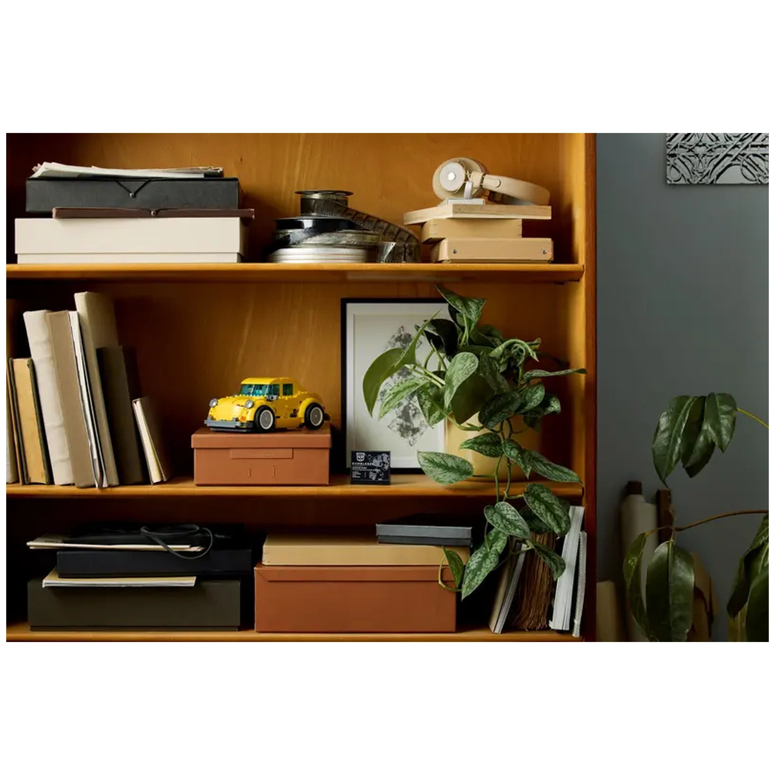 LEGO Bumblebee construction set in yellow, displayed on a wooden shelf among books, boxes, and a potted plant, with framed art in the background.