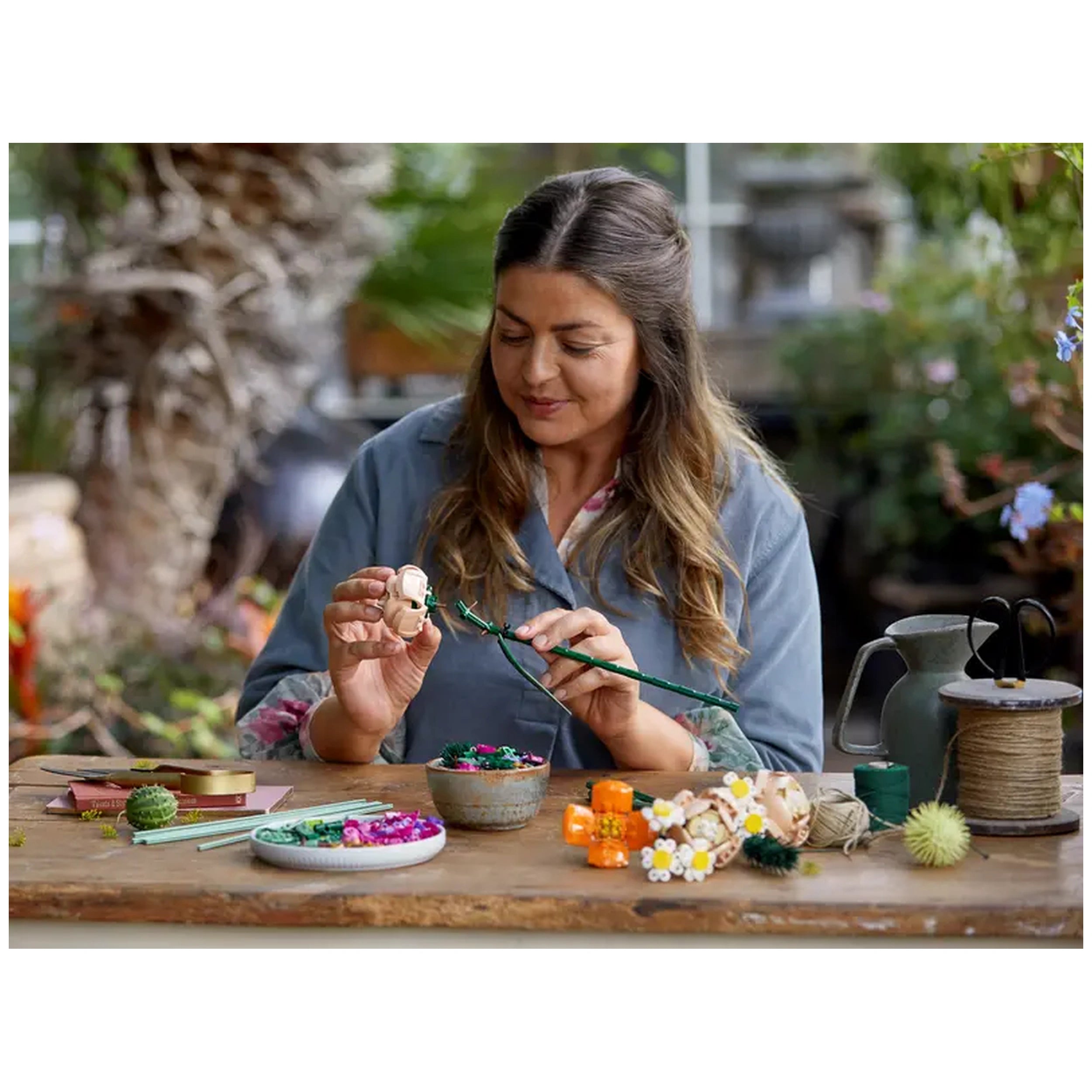 A woman with long, brown hair is carefully crafting a flower arrangement outdoors, surrounded by various colorful artificial flowers and tools on a wooden table. She holds a flexible green stem and appears focused on her creative project, with vibrant pots and a rustic watering can in the background.