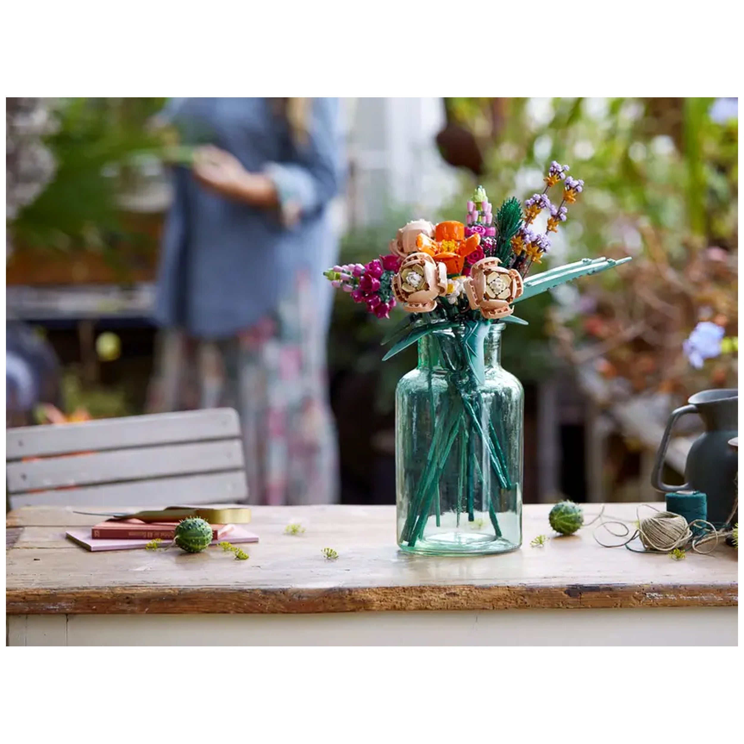 Colorful LEGO Flower Bouquet arranged in a clear glass vase on a wooden table, with craft materials and a blurred background of greenery.