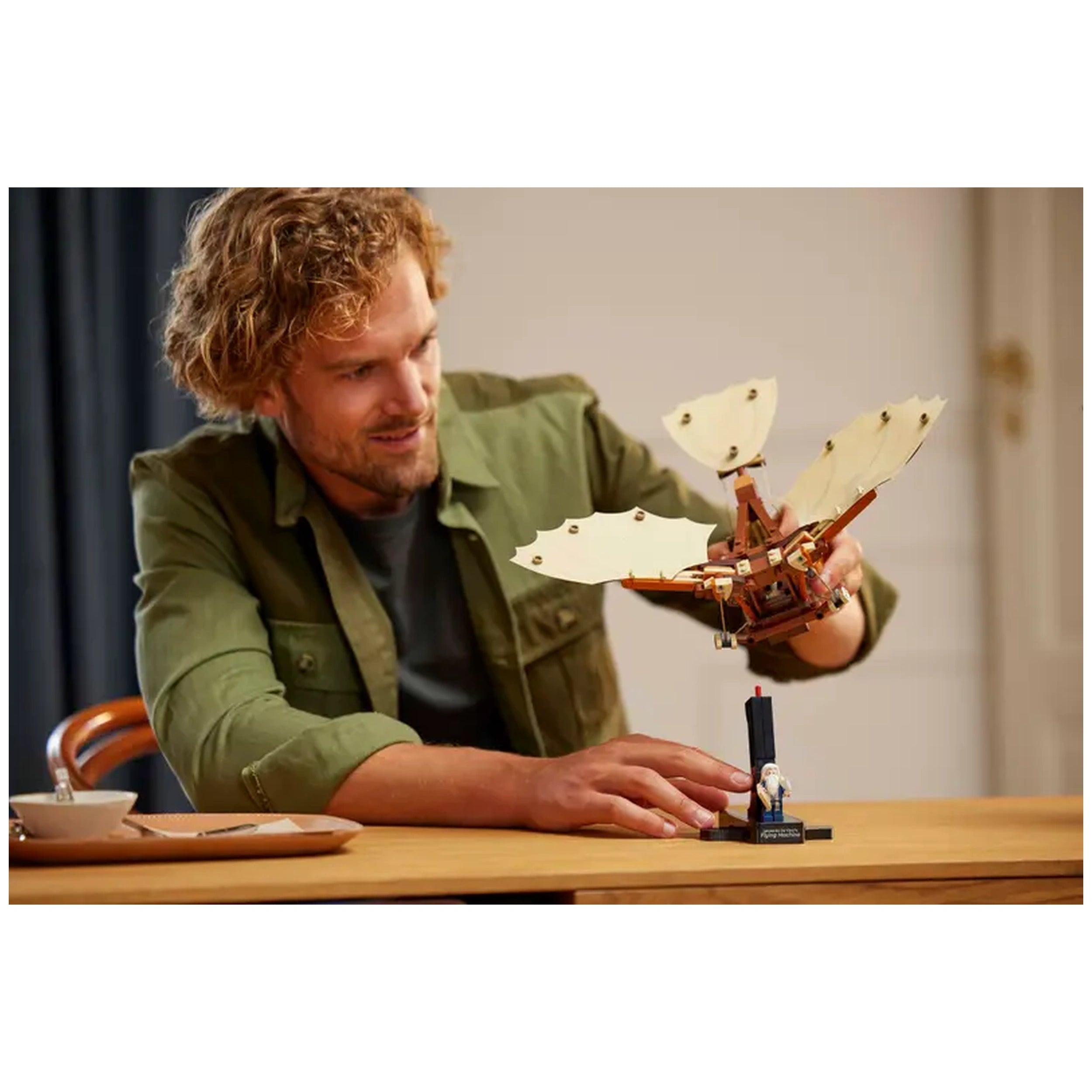 Adult male with curly hair smiles while assembling LEGO® Icons Leonardo da Vinci's Flying Machine on a table, showcasing intricate flappable wings.