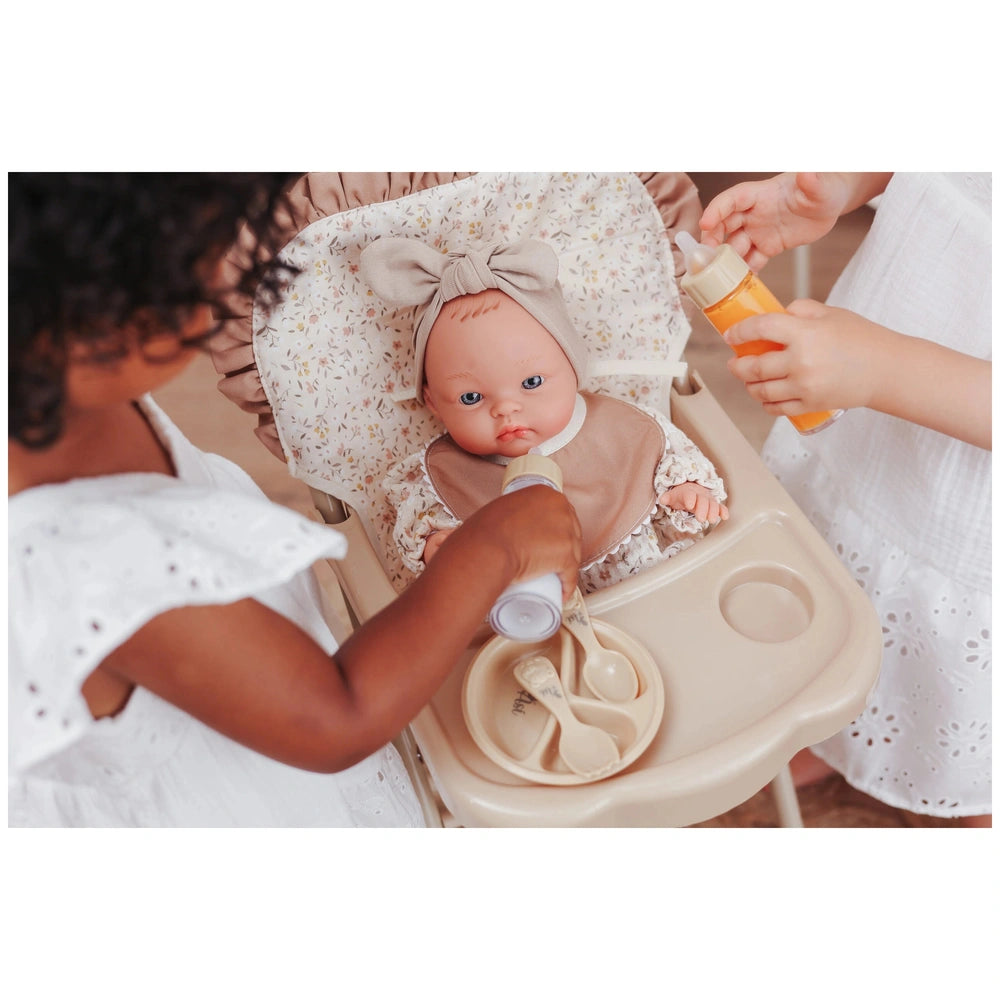 ASI Dolls high chair with tray and bib, featuring a baby doll in a beige outfit being fed in a domestic nursery setting.