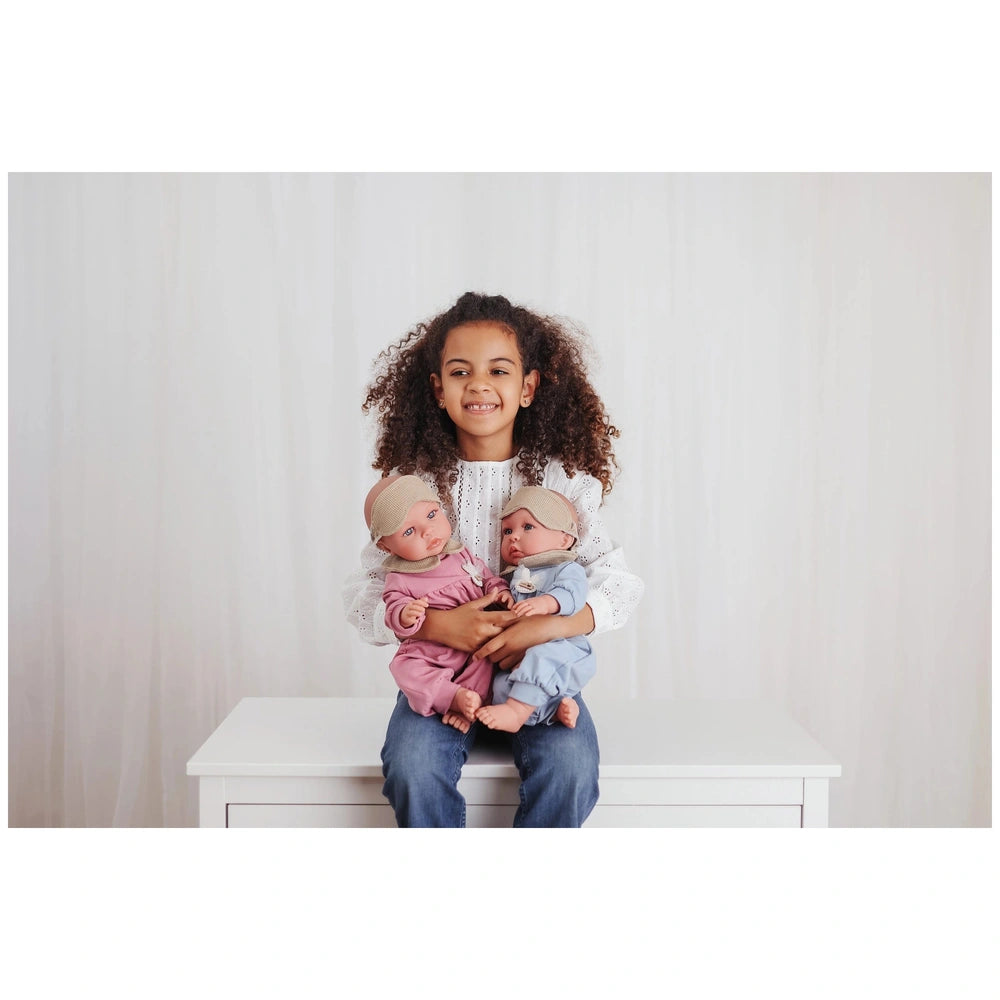 Child holding two realistic dolls, one in pink pajamas, one in blue pajamas, both wearing headbands, seated on a white table.