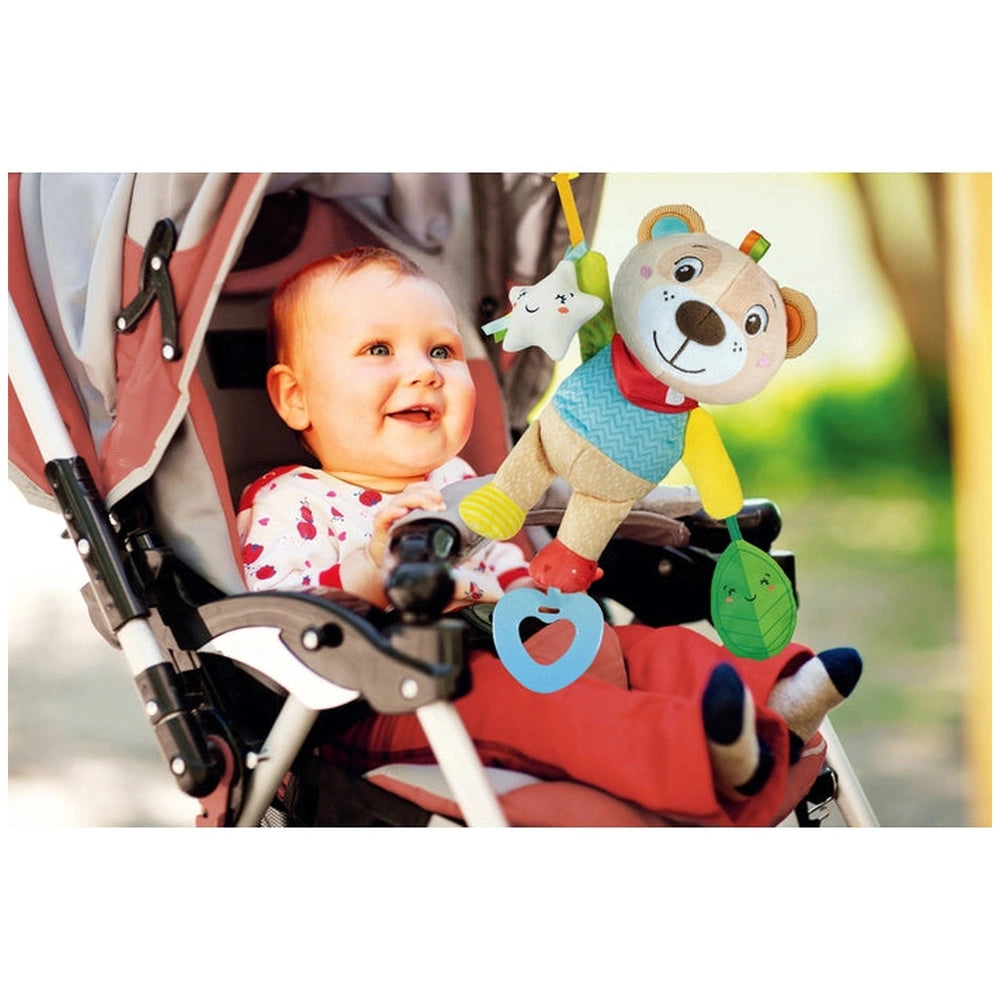 Baby Clementoni soft bear stuffed animal is held by a smiling child in a stroller, surrounded by greenery outdoors.