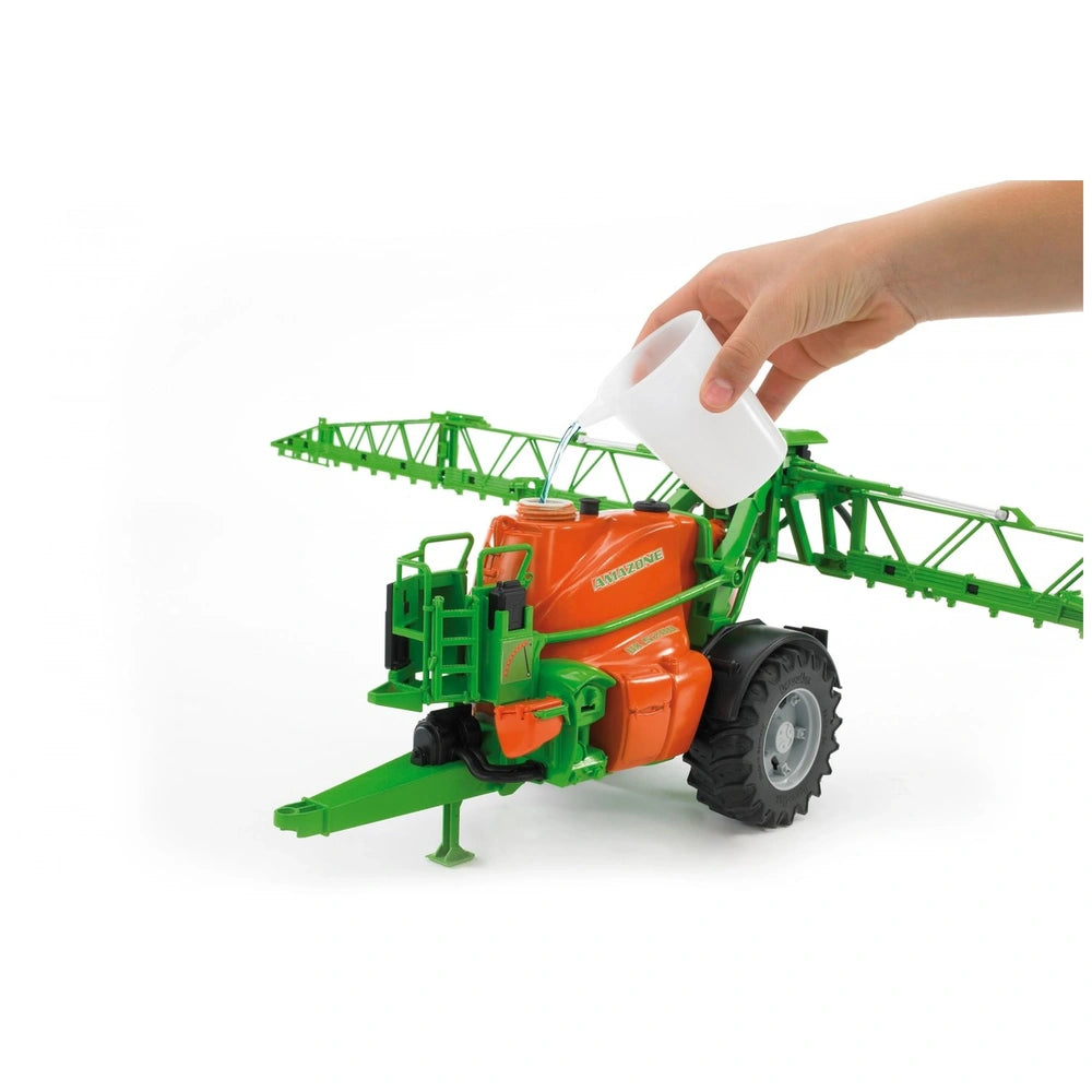 A hand pours water into an orange and green Bruder Amazone UX 5200 toy field sprayer with extendable booms.