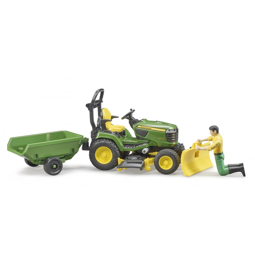 Green John Deere lawn tractor with yellow seat, attached trailer, and figure kneeling to attach a yellow snow plough.