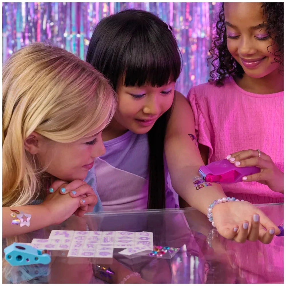 Cool Maker Shimmer Me Body Art Set displayed on a clear table with three curious young girls in colorful clothing.