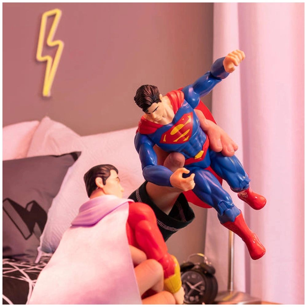 Superman action figure in a superhero pose on a bed with a pink bedspread, alongside a smaller red-costumed figure.