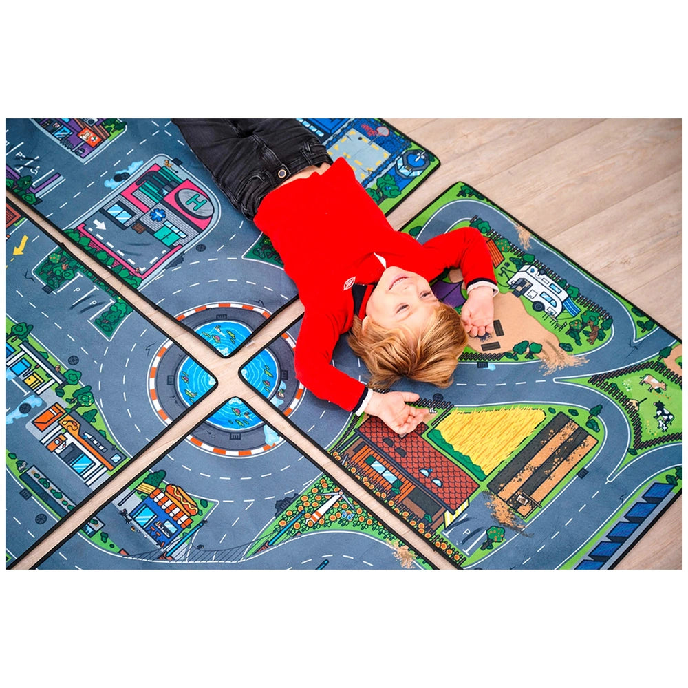 Child in a red shirt and black pants lies on a fabric playmat with a printed road map design, engaging with play vehicles.