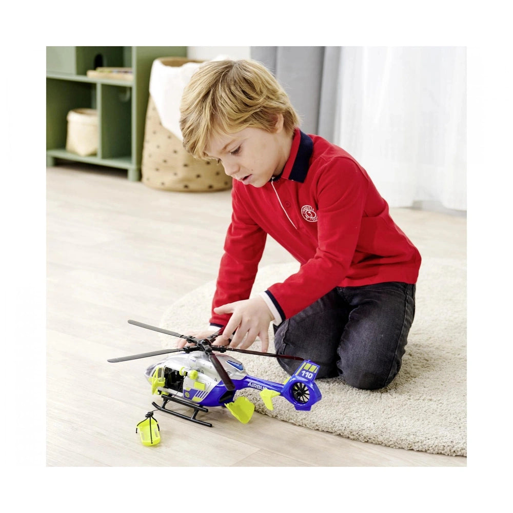 Boy with blonde hair plays with a blue and yellow Dickie police helicopter on light wood flooring and a beige rug.