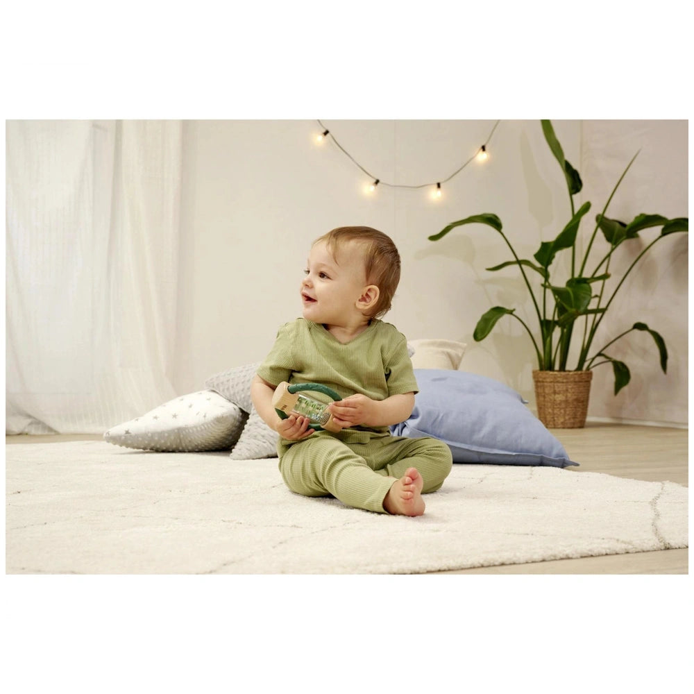 Eichhorn Baby HiPP Sound Rattle is held by a smiling child in a green outfit, seated on a white rug with a plant nearby.