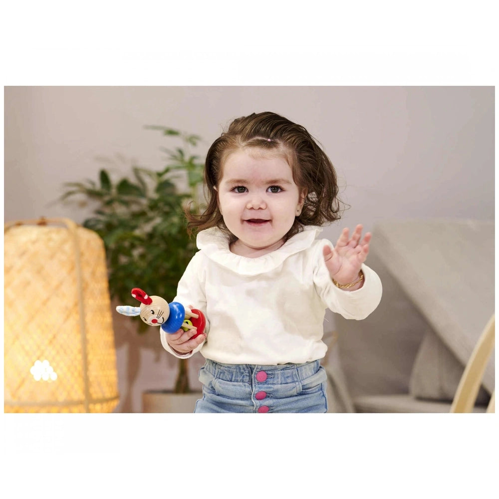 Baby wooden rattle rabbit held by a smiling toddler in a cozy indoor setting with a plant in the background.