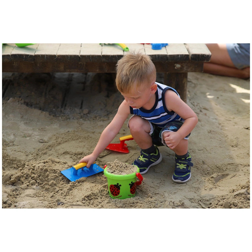 Gowi beach bucket in green and red, alongside blue and yellow shovels, as a child plays in the sand.