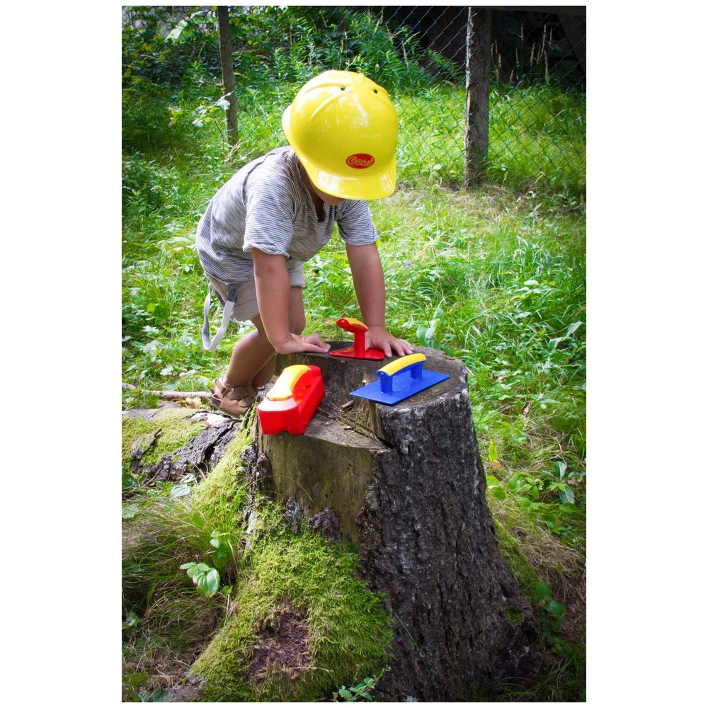 Gowi Design Bricklaying Set features a person in a yellow hard hat holding a red and yellow tool near a mossy stump and wire 
