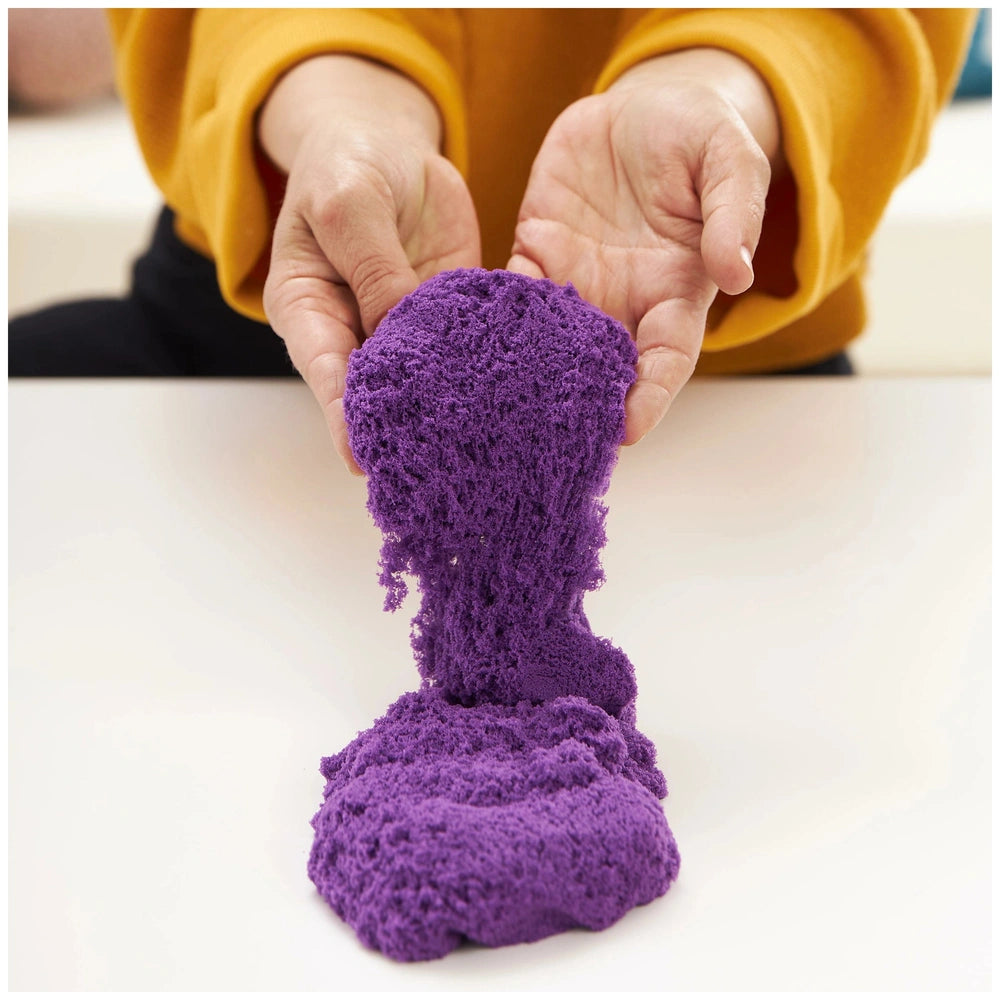 Kinetic Sand Sandbox Set features a person's hands holding a fluffy purple substance in an indoor setting.