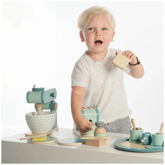 Child with blonde hair in a white t-shirt plays with a wooden block in front of a blue toy kitchen with a white countertop.