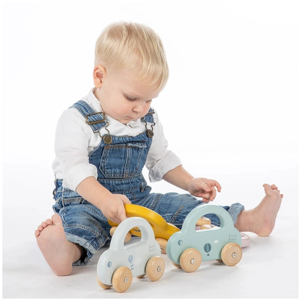 Little Car push and pull toy held by a child in blue denim overalls, sitting on a white surface with wooden-wheeled cars.