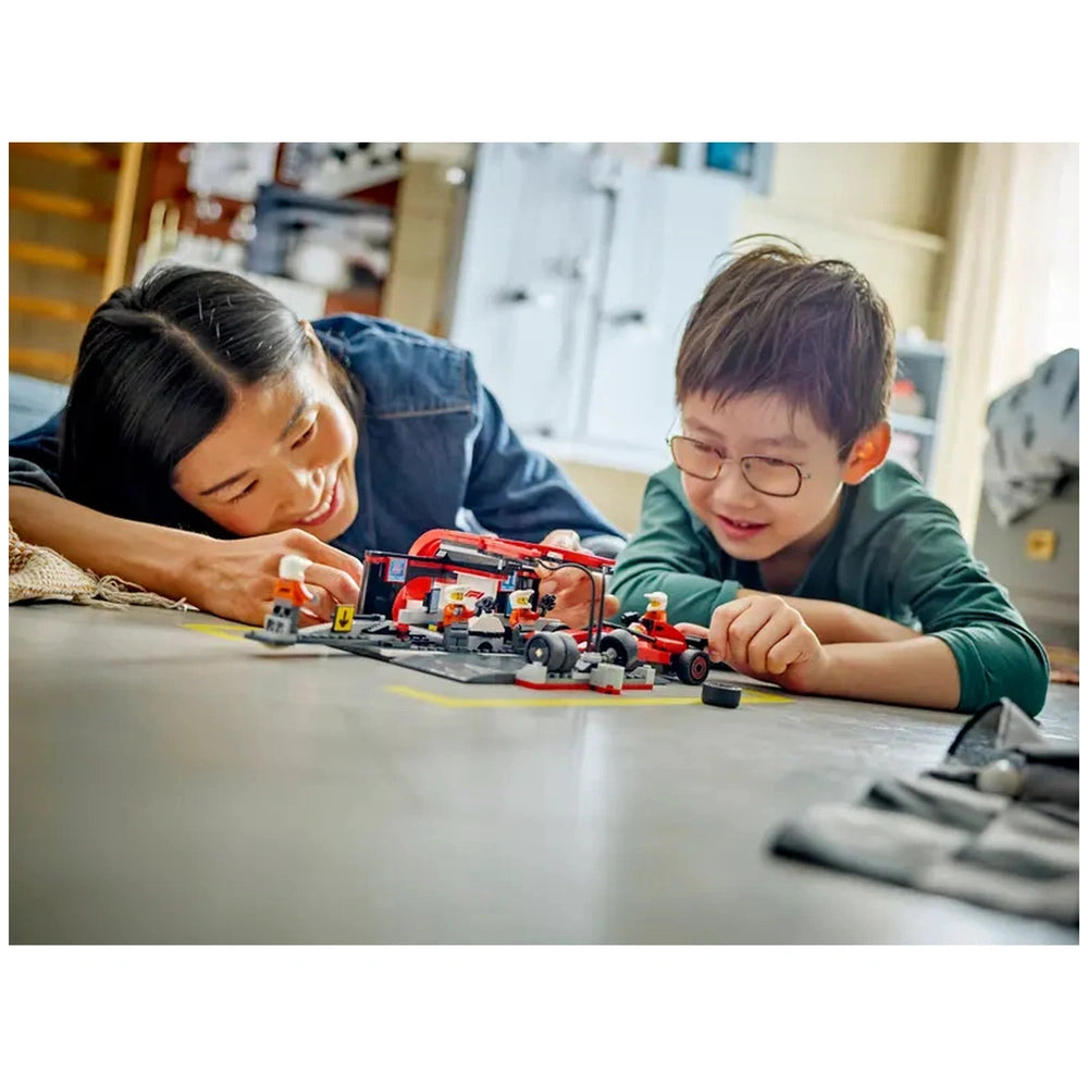 A smiling woman and a young boy engage in building the LEGO City F1 Pit Stop set on a floor. The scene features a colorful Ferrari car, pit crew minifigures, and accessories, creating a lively pit stop atmosphere.