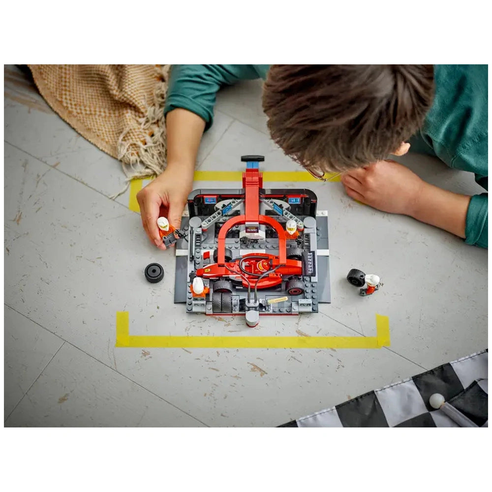 A child plays with the LEGO City F1 Pit Stop set on a light-colored floor, assembling a vibrant red Ferrari car and four pit crew minifigures, surrounded by tools and tires.