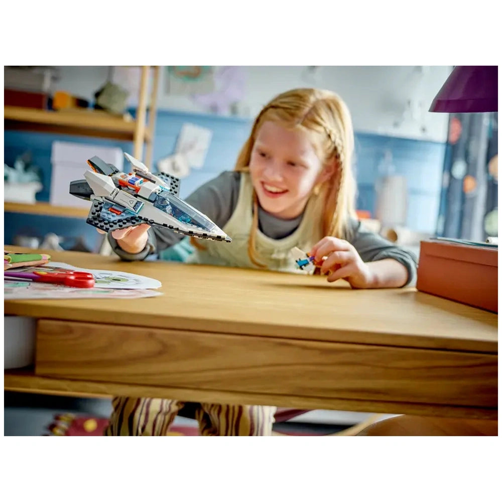 A child with long, blonde hair smiles while holding a LEGO® spaceship and a minifigure at a wooden table, surrounded by colorful craft materials.