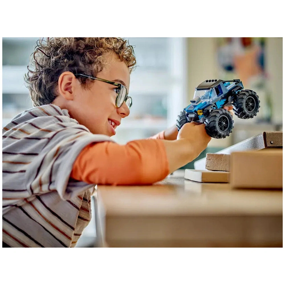 A smiling child with curly hair and glasses is playing with a blue LEGO® monster truck, engaged in imaginative play at a wooden table.