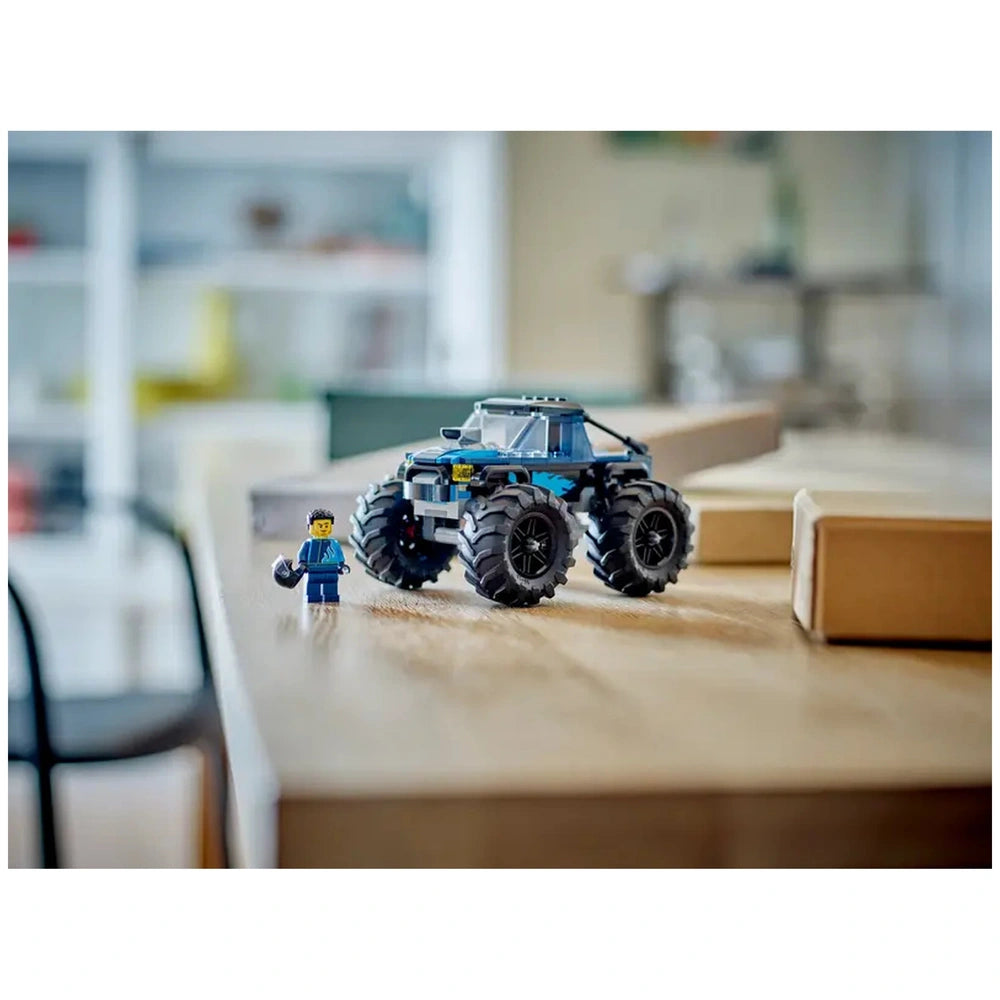 LEGO® City Blue Monster Truck with oversized tires on a wooden table, next to a driver minifigure holding a helmet.