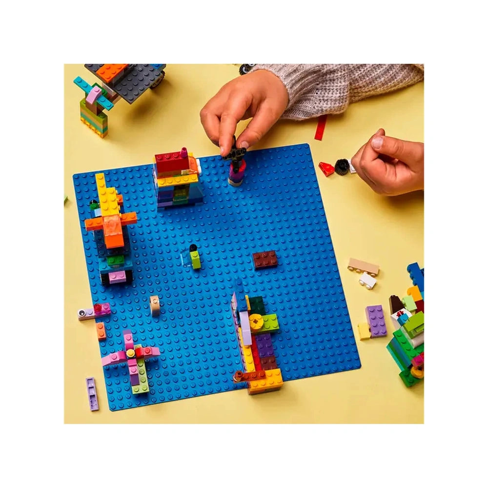 Colorful LEGO constructions scattered across a vibrant blue baseplate, with hands actively building and arranging pieces.