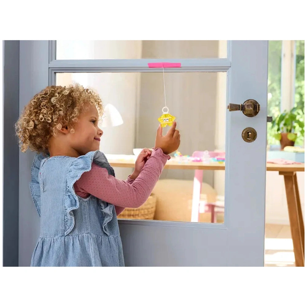 A young girl with curly hair smiles while holding a colorful hanging toy near a transparent door. She wears a blue dress with ruffles and a pink long-sleeve shirt. A bright, spacious room is visible in the background.