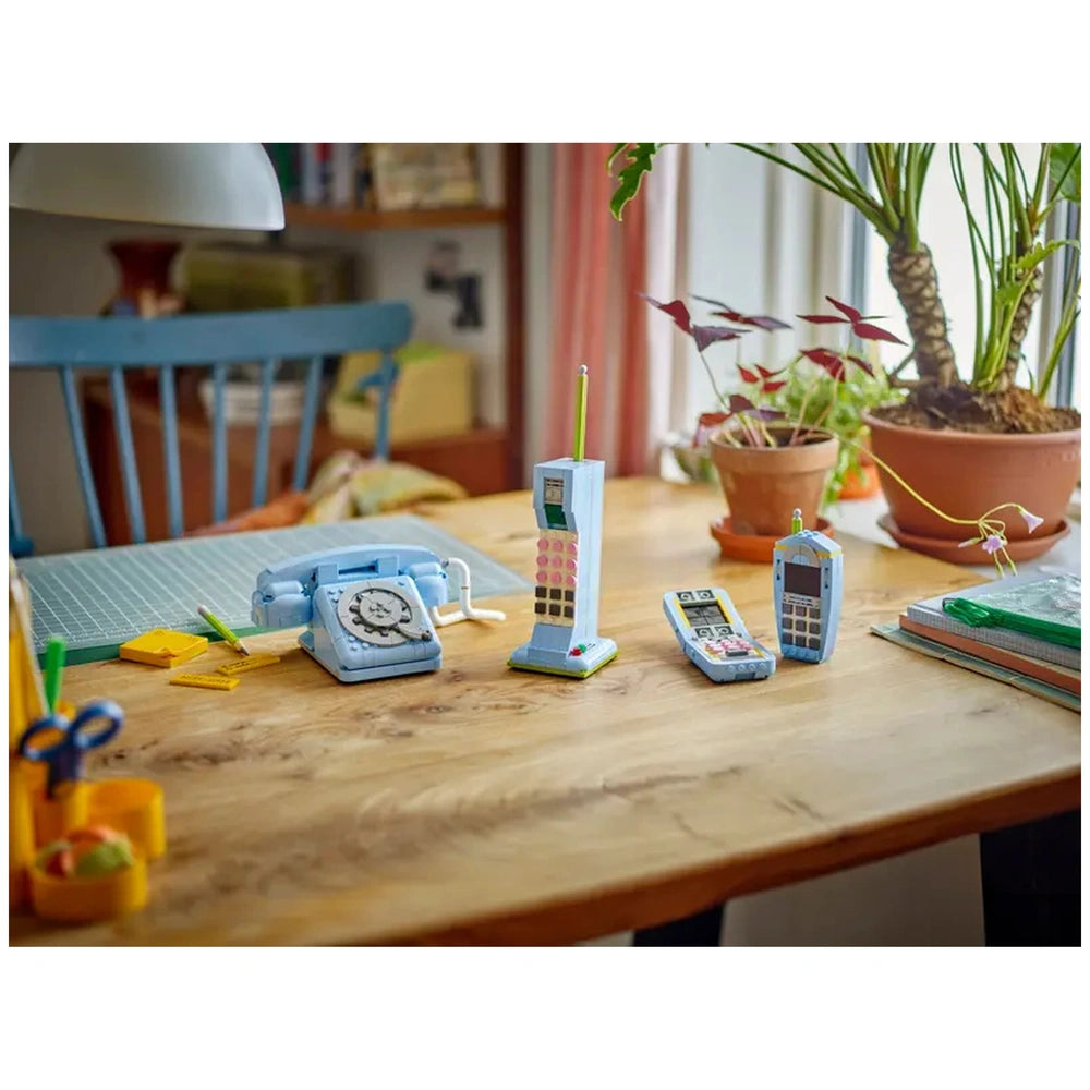 Retro telephone and two modern toy phones displayed on a wooden table, surrounded by colorful stationary and a potted plant, capturing a nostalgic vibe.