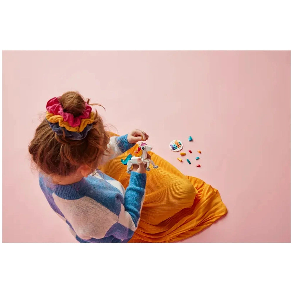 A young child with curly hair, wearing a blue and white sweater, holds a colorful LEGO unicorn construction set. The child sits on a vibrant yellow skirt on a pink background, surrounded by scattered LEGO pieces.