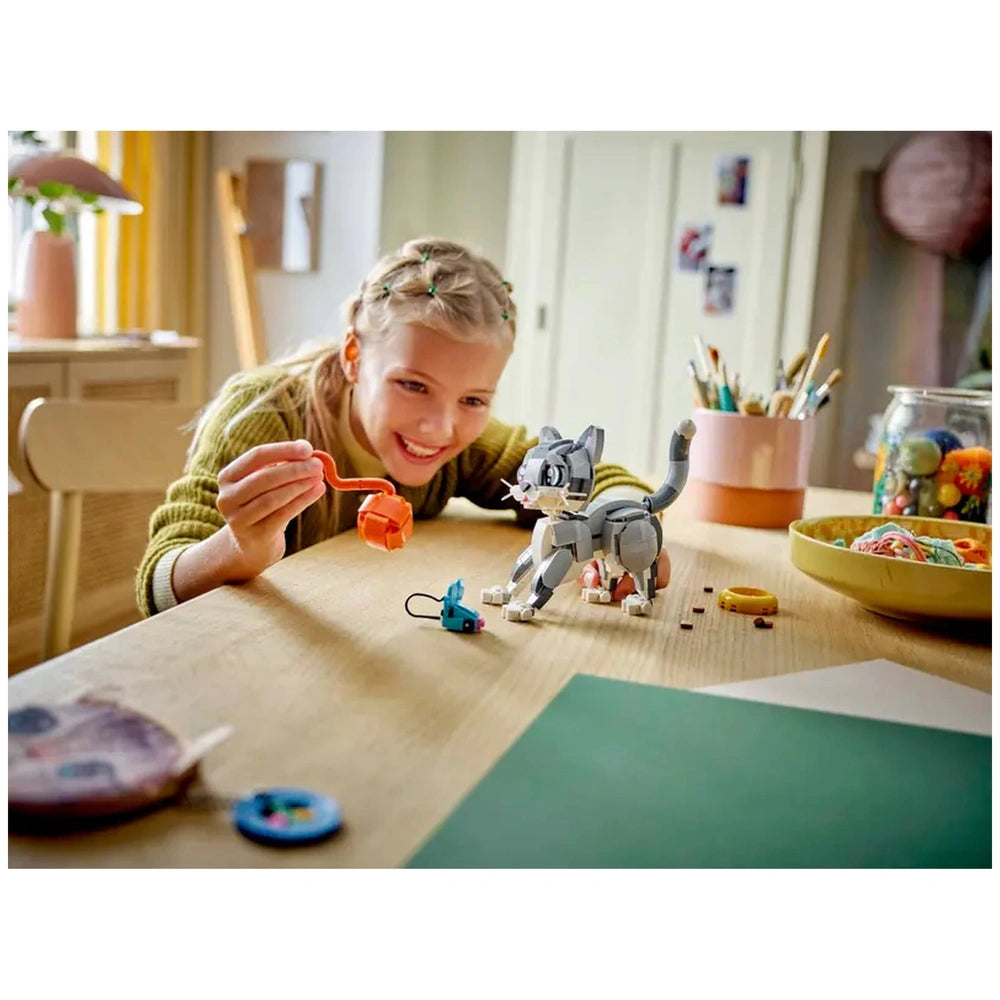 A young girl smiles while playing with the LEGO Creator 3in1 Playful Cat set on a wooden table. She holds an orange toy while the gray cat figure stands nearby, accompanied by a blue toy mouse and colorful accessories. Bright natural light fills the room, enhancing the playful atmosphere.
