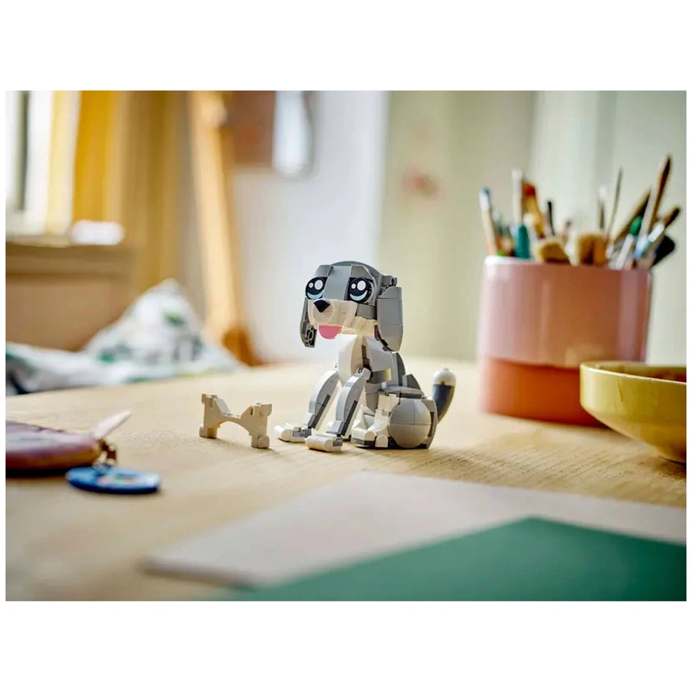 LEGO Creator 3in1 Playful Cat construction set showcasing a grey and white cat with expressive eyes, sitting on a wooden table with a toy bone nearby, surrounded by art supplies and a bowl.