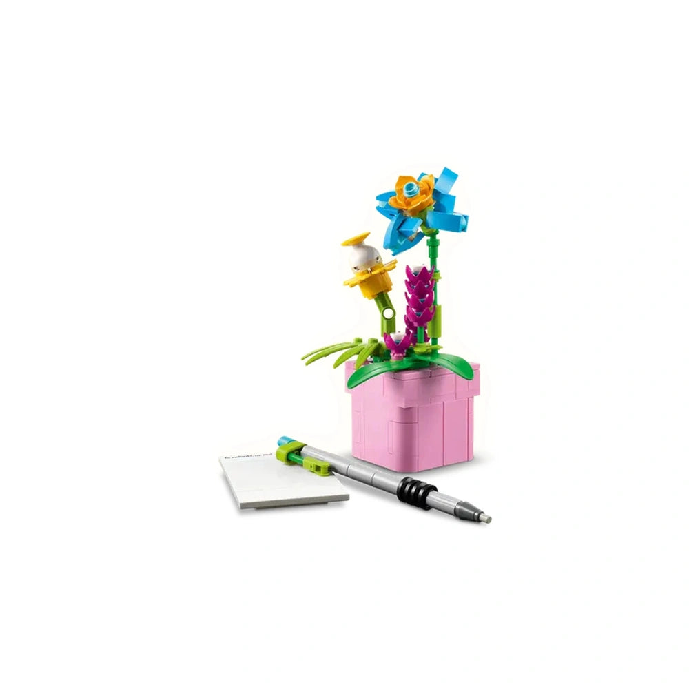 Colorful LEGO® flowers in a pink pot, featuring blue, yellow, and purple blooms, alongside a white sheet of paper and a silver pen.