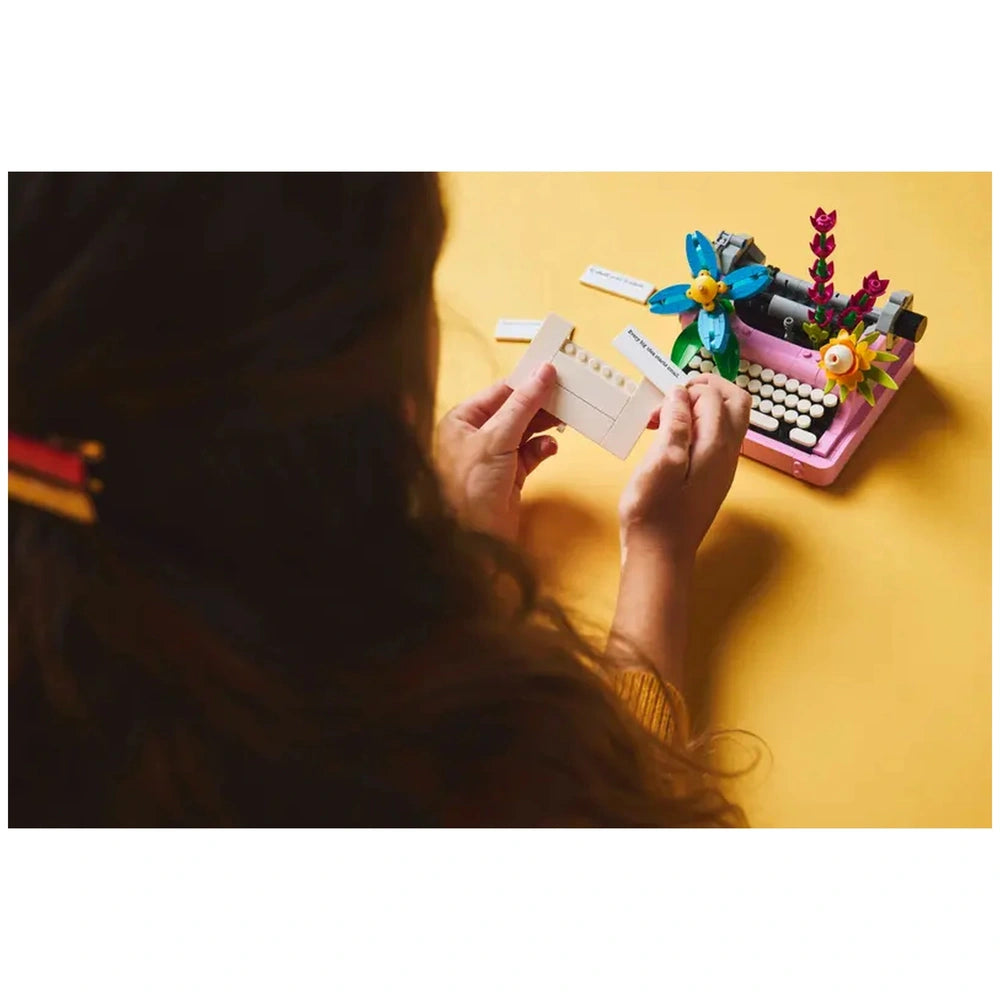 A child holds a brick-built sheet of paper in front of a colorful LEGO® Typewriter, featuring vibrant flowers on a bright yellow background.