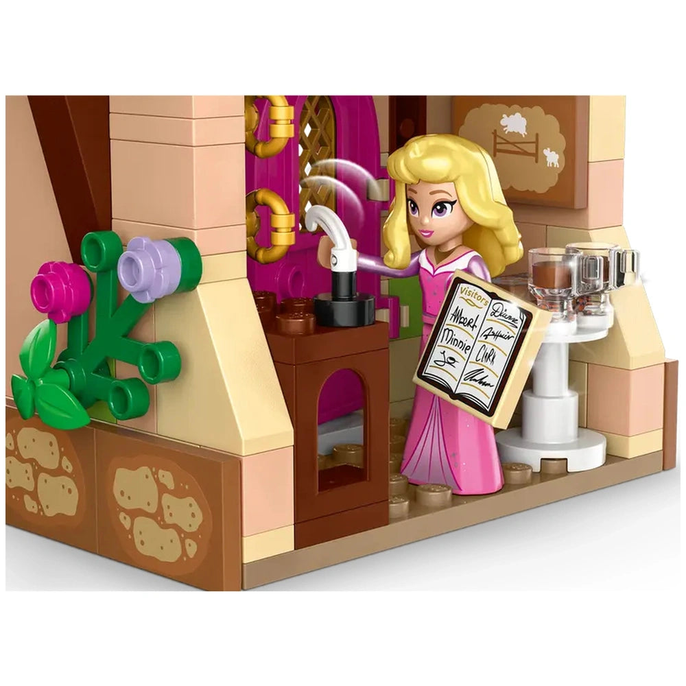 Aurora, wearing a pink dress, stands inside a LEGO® café, holding a visitor book while using a coffee maker. Colorful flowers and decorative elements adorn the walls.