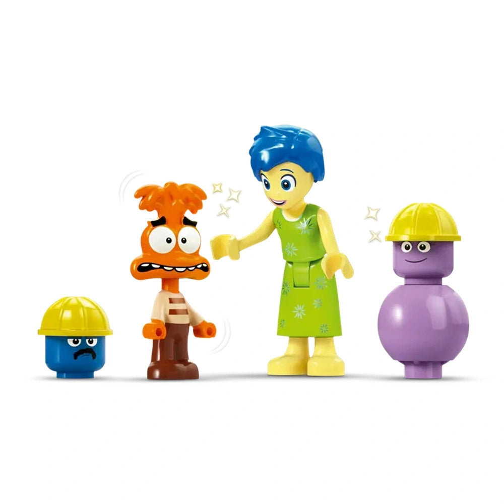 Joy and Anxiety mini-doll figures interact, with Joy in a green dress and blue hair comforting anxious Anxiety. Two purple and blue mind workers with hard hats stand nearby, adding to the scene.