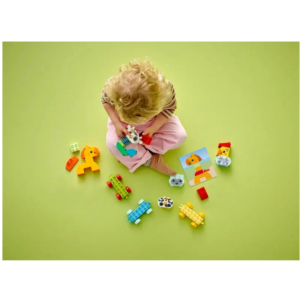 A child with curly hair sits on a green mat, playing with colorful LEGO DUPLO animal figures and vehicles. Brightly colored blocks are scattered around, including a buildable elephant, train carriages, and an instruction card featuring an animal. The child is focused on assembling a piece of the animal train, showcasing imaginative play.