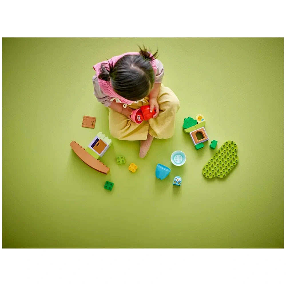 A toddler sits on a green floor, focused on a red toy while surrounded by colorful LEGO® DUPLO® blocks and pieces, including an adorable owl and a tree house.