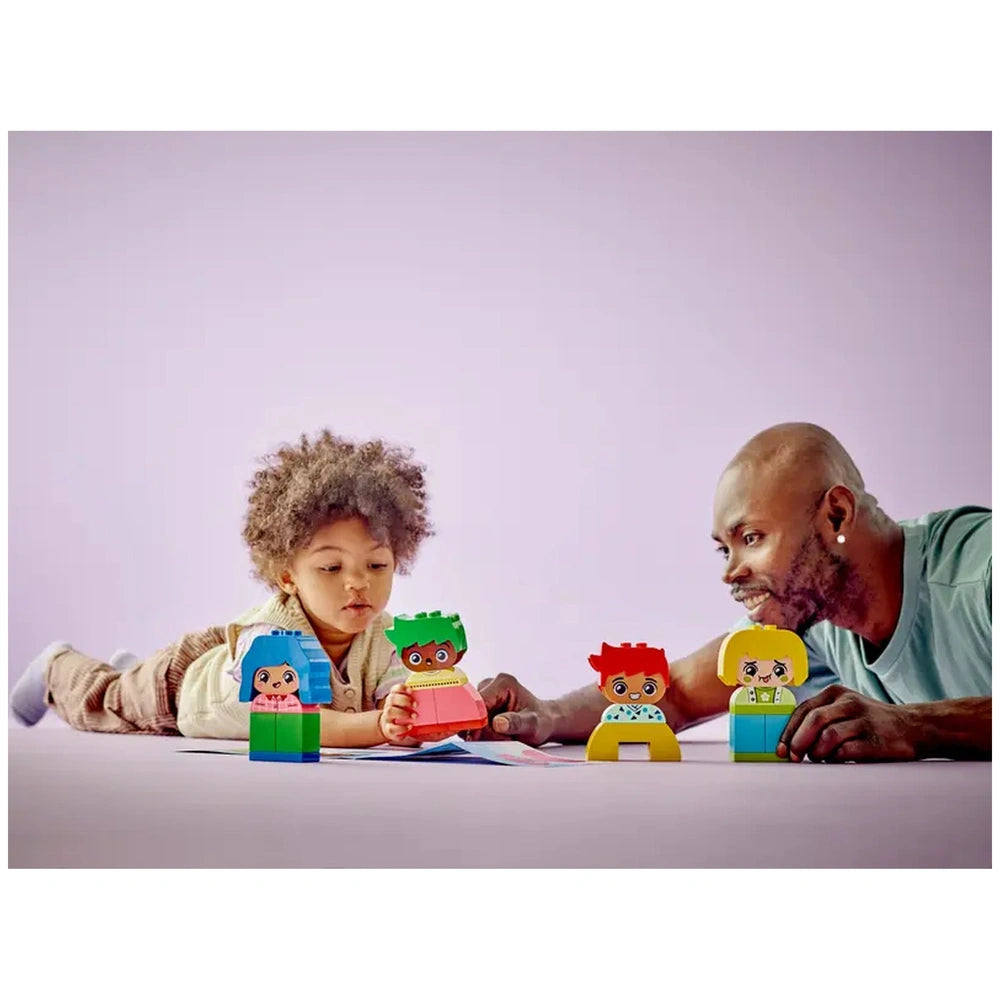 A toddler and an adult play together on a soft surface, engaging with colorful LEGO DUPLO characters that express emotions. The background is soft purple, enhancing the joyful learning atmosphere.