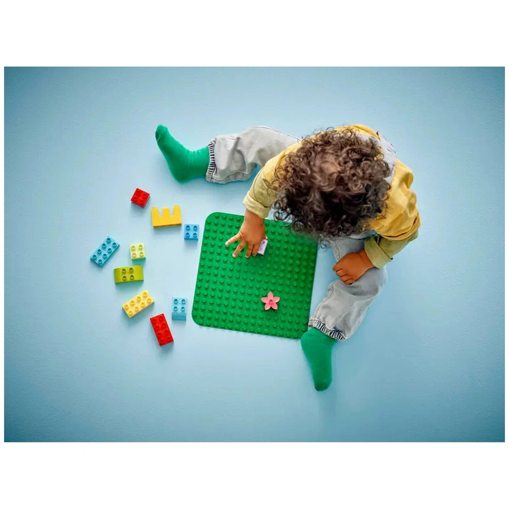 A young child sits on a light blue surface, placing a tile on a green LEGO® DUPLO® building plate. Surrounding the child are colorful building bricks in red, yellow, blue, and green, indicating creative playtime.