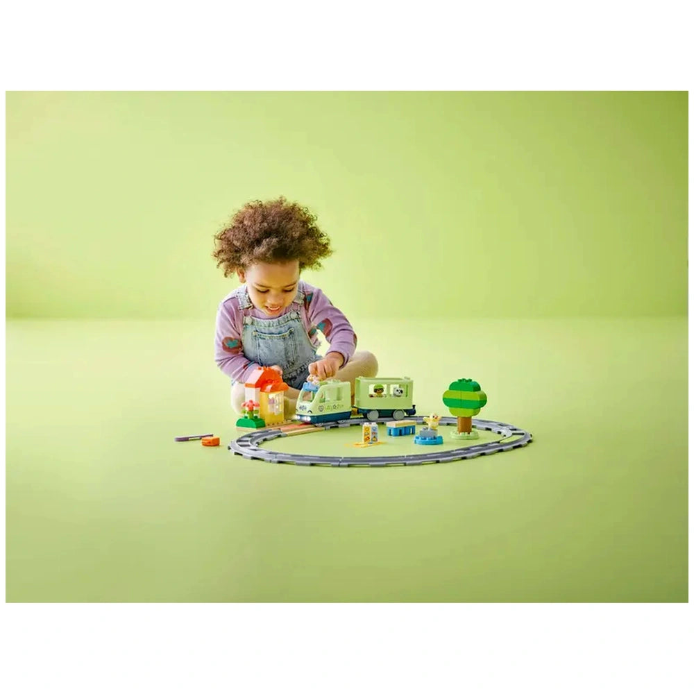 A toddler with curly hair sits on a light green floor, assembling the LEGO® DUPLO® Interactive Adventure Train set, which includes a train, buildings, and a tree.