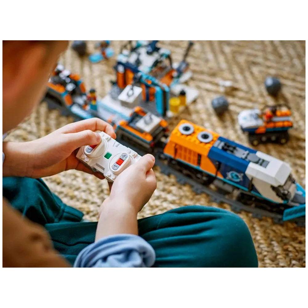 A child holds a white remote control, focused on a colorful LEGO Arctic train set with various cars and an outpost on a textured carpet.