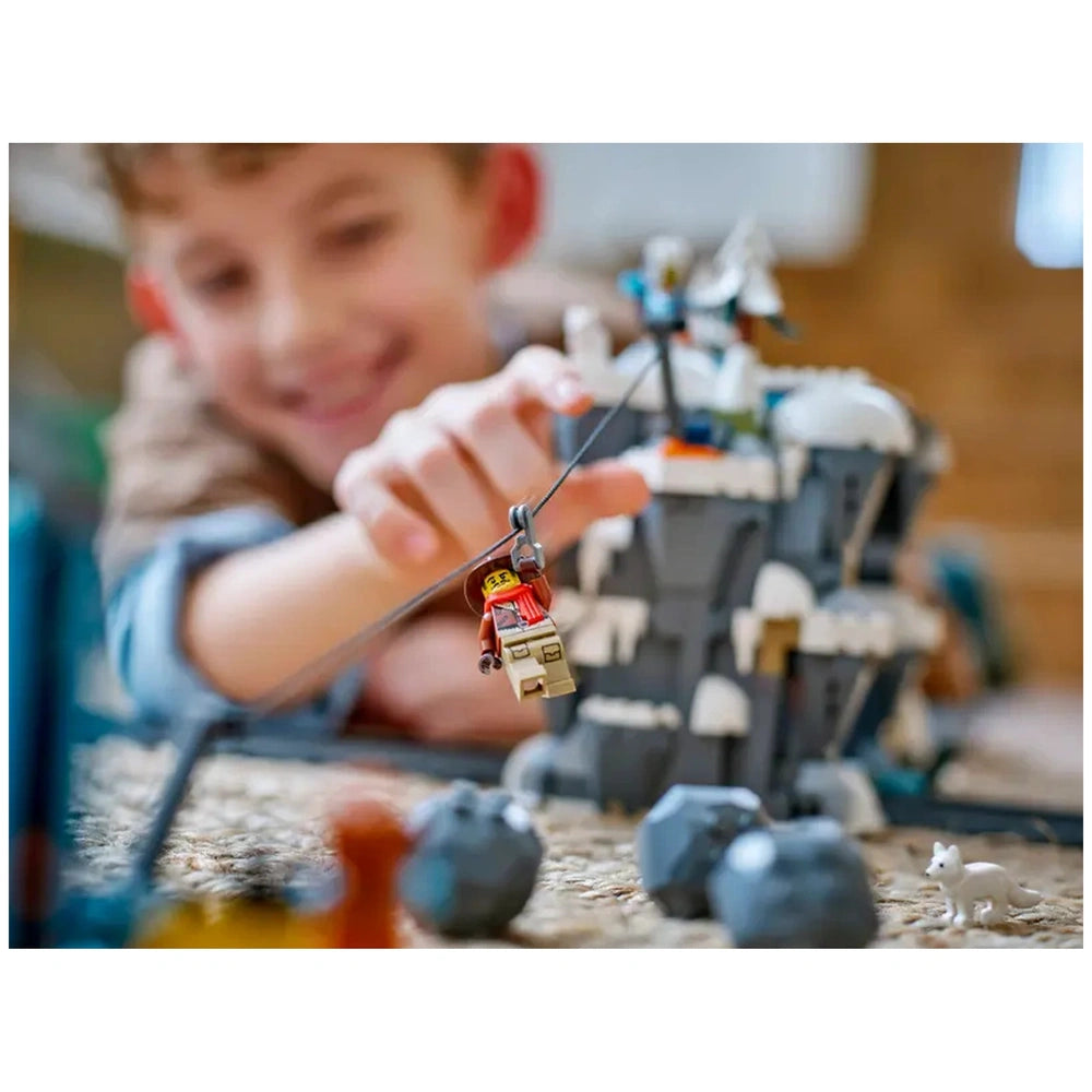 A child joyfully interacts with a LEGO explorer figure hanging from a cable, set against a snowy mountain backdrop filled with rocks and animals.